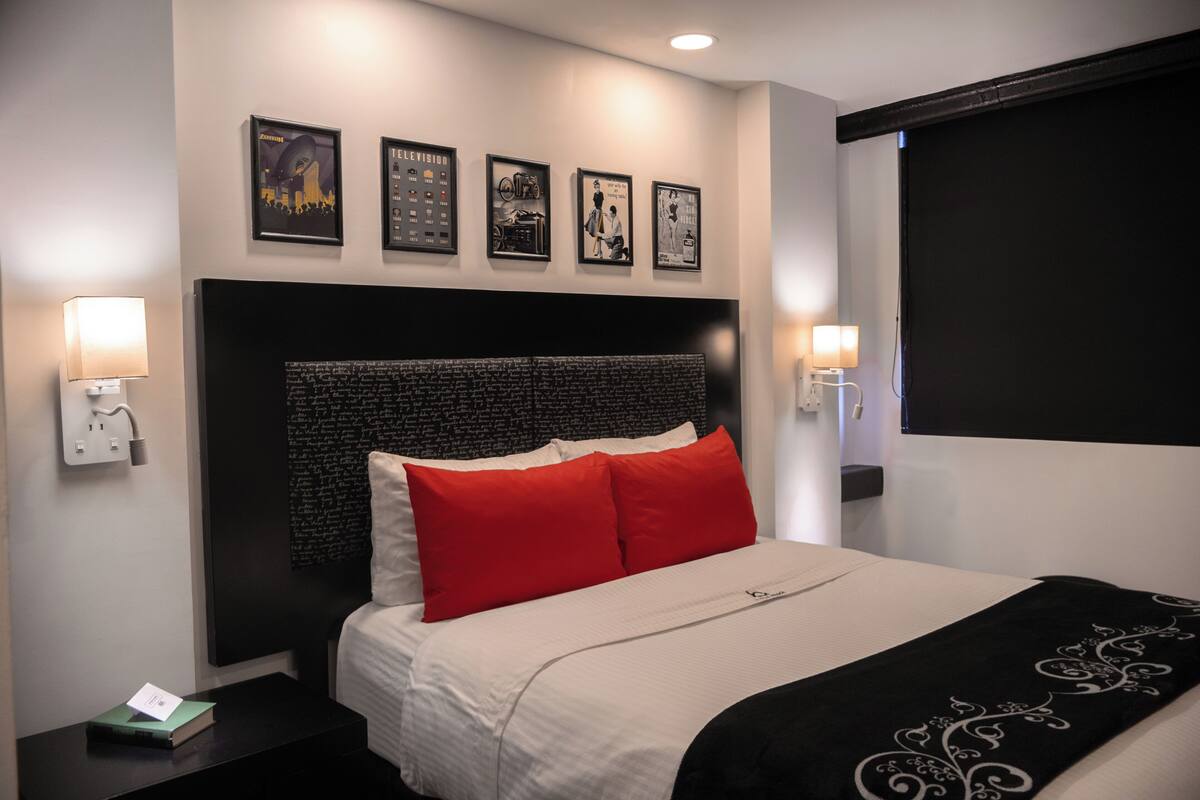 Habitación de hotel con una cama grande, cabecera negra con texto, almohadas rojas y blancas, dos lámparas de noche y cuadros en la pared.