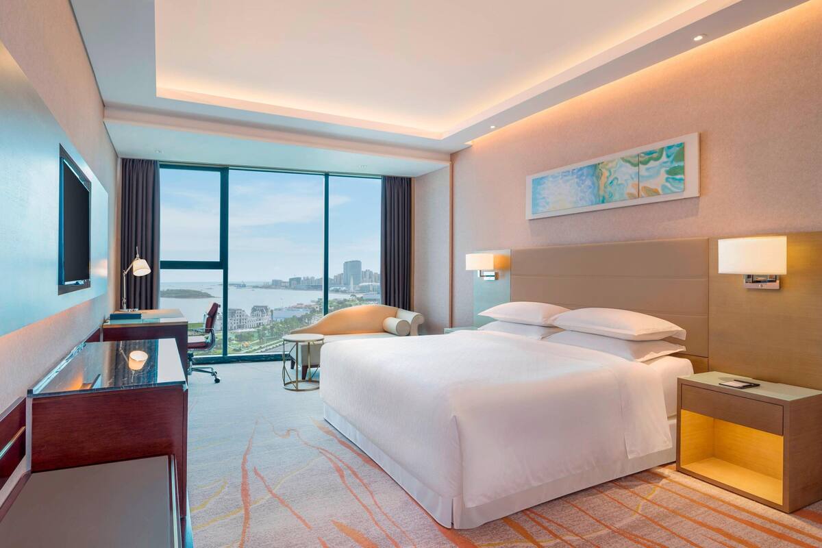 Een hotelkamer met een groot bed, een bureau, een stoel, een nachtkastje en uitzicht op de stad door grote ramen.