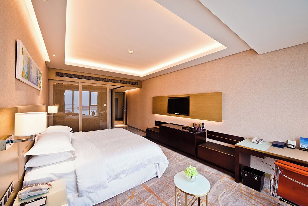 A modern hotel room with a large bed, a desk, a TV, and a bathroom with a glass door.