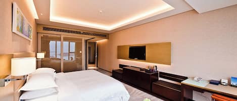 A modern hotel room with a large bed, a desk, a TV, and a bathroom with a glass door.