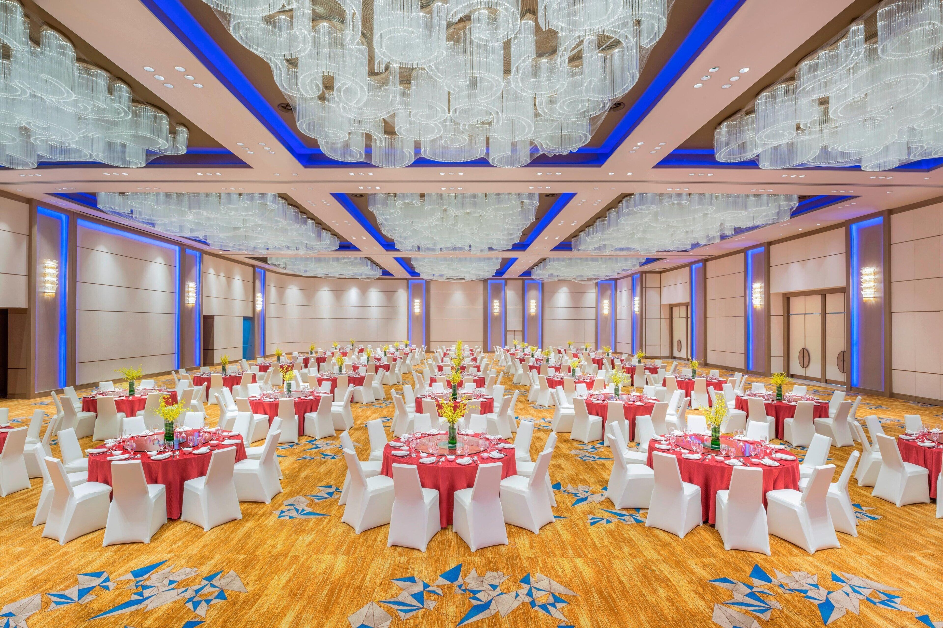 A large banquet hall with rows of round tables, each set with white chairs and red tablecloths, featuring a wooden floor and modern chandeliers.