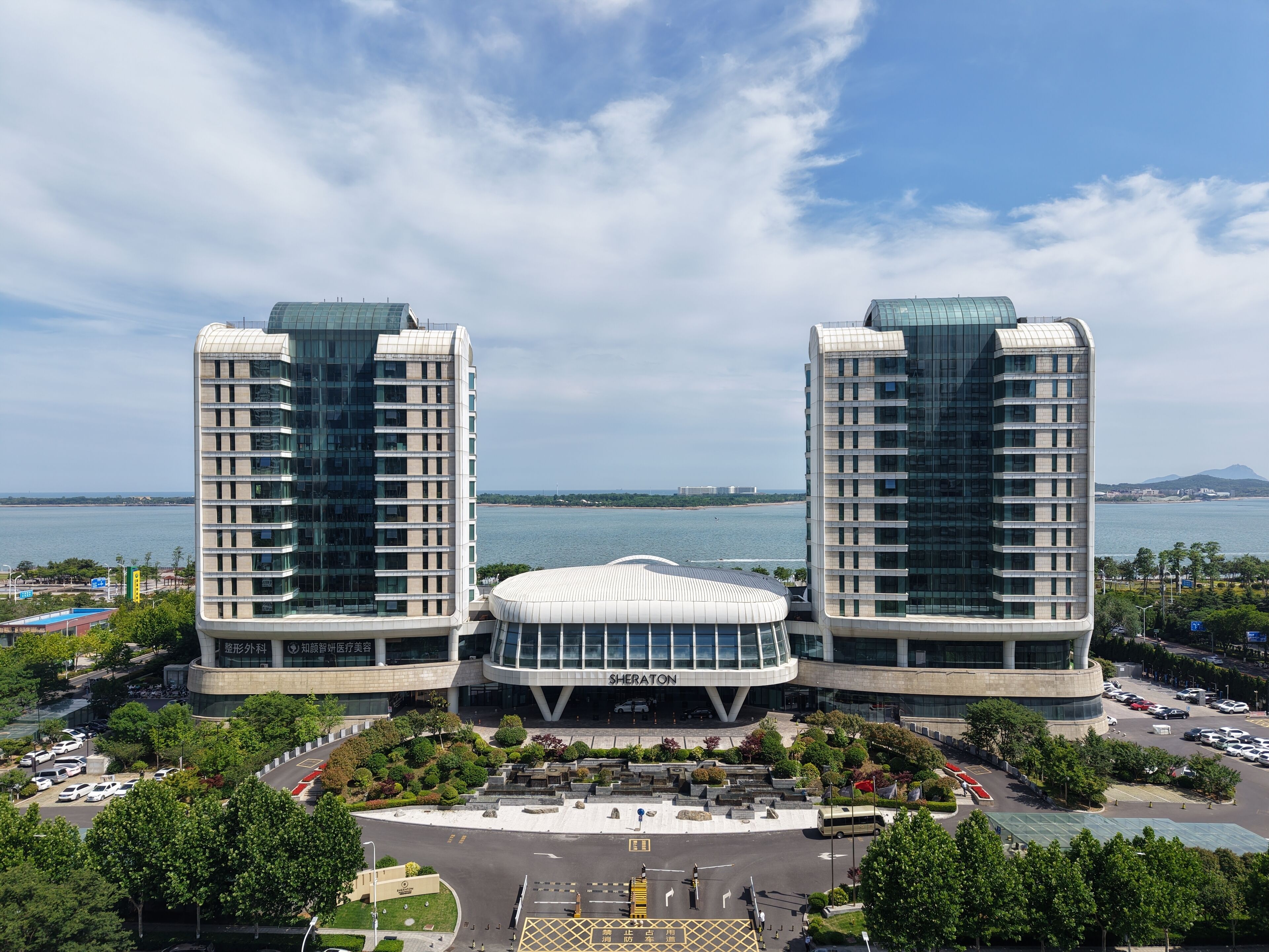 A modern building complex with two tall towers and a central dome structure, surrounded by greenery and a parking area.