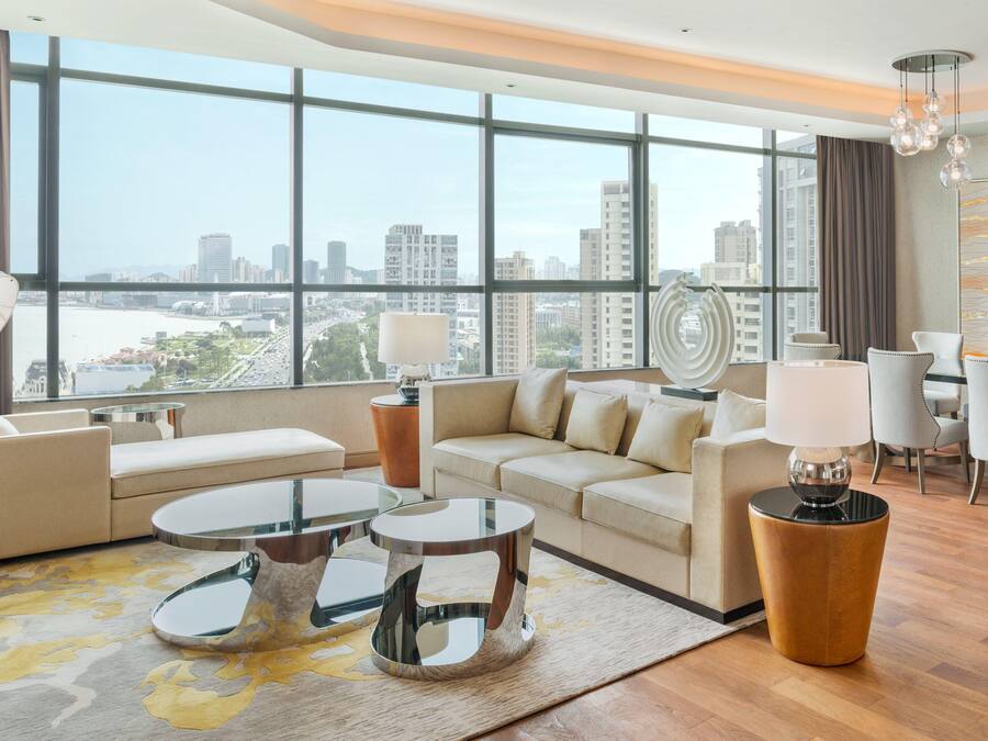 A modern hotel room with a large window offering a city view, a sofa, a coffee table, and a dining area with chairs and a lamp.