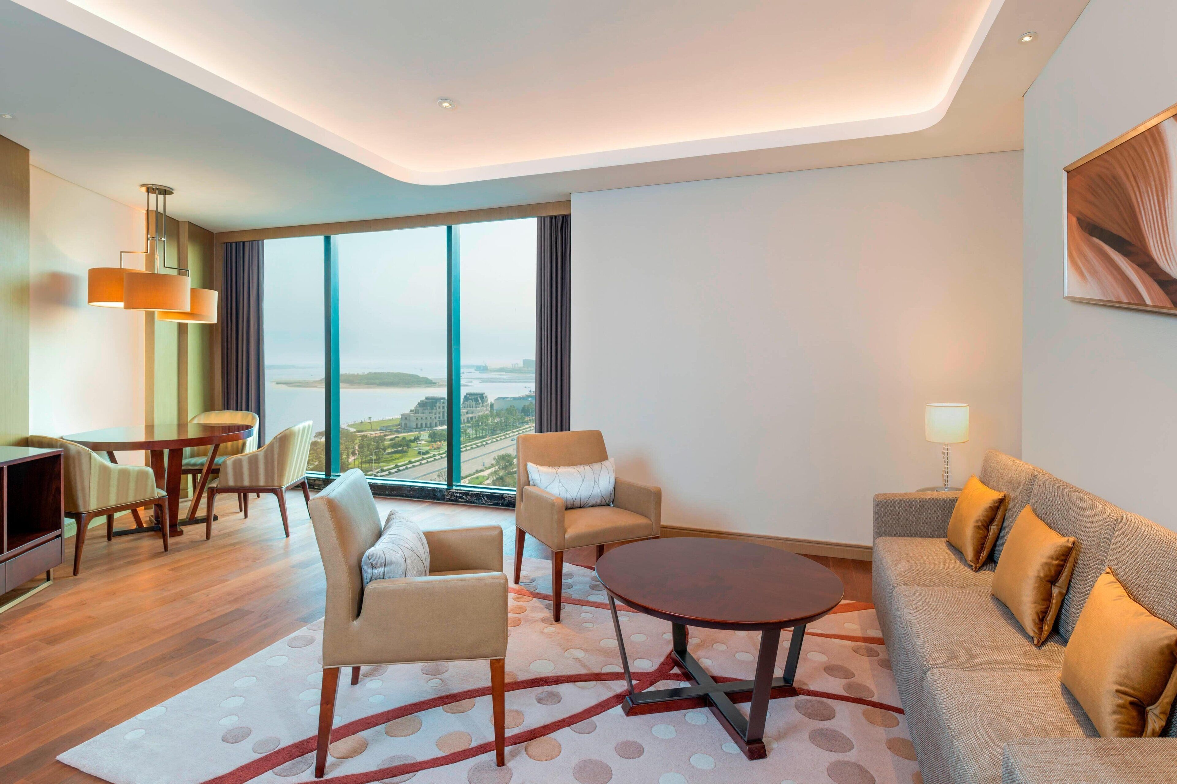 A modern hotel room with a large window, a sofa, a dining table, and a coffee table.