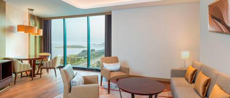 A modern hotel room with a large window, a sofa, a dining table, and a coffee table.