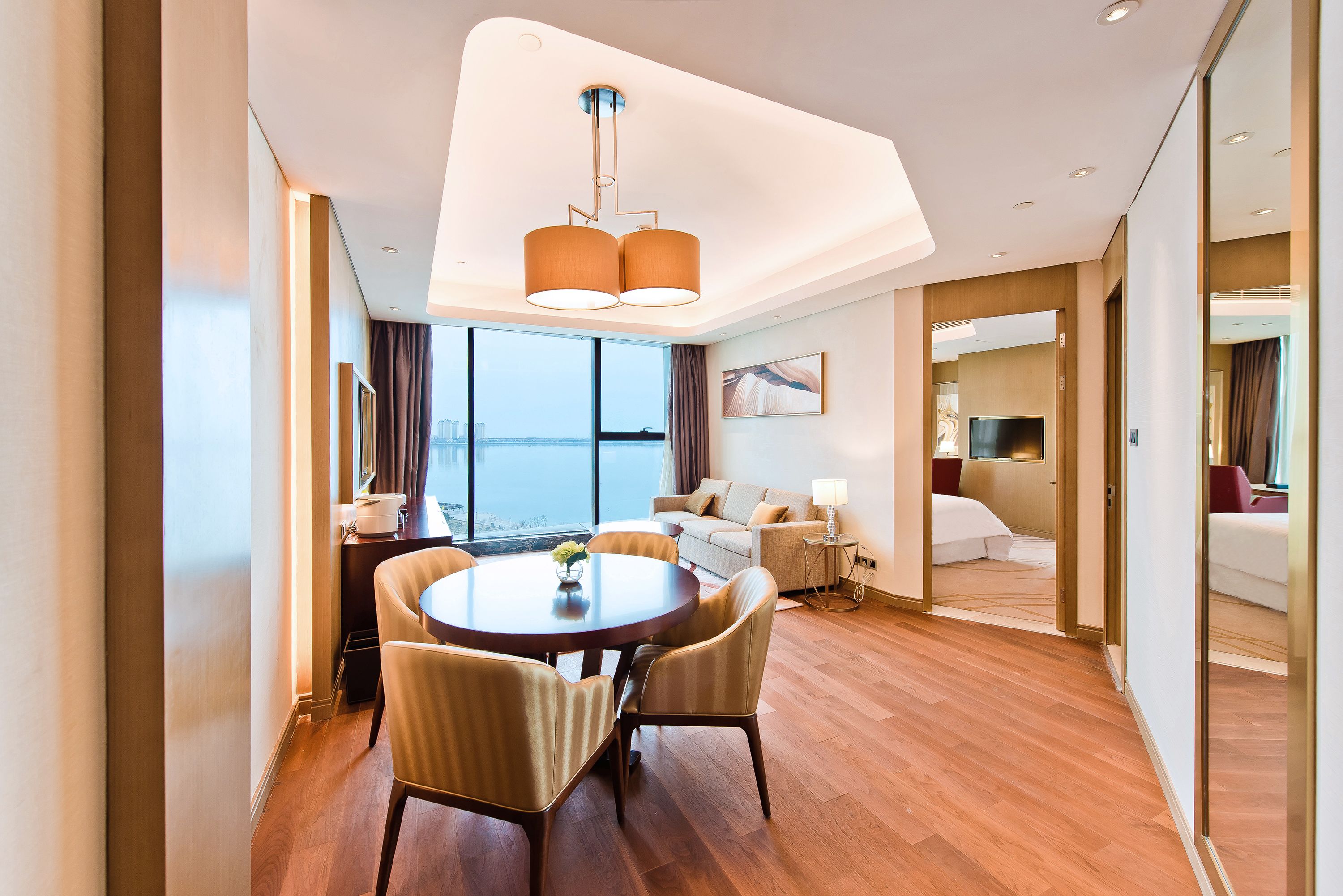 A modern hotel room with a dining area, a sofa, a TV, and a large window with a view.