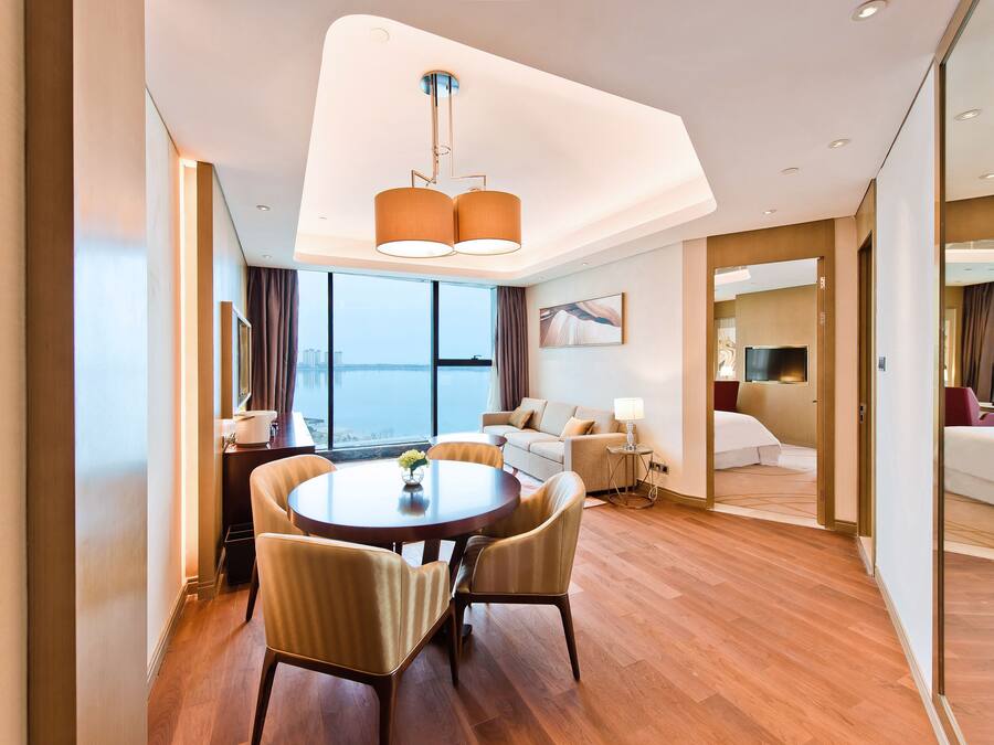 A modern hotel room with a dining area, a sofa, a TV, and a large window with a view.
