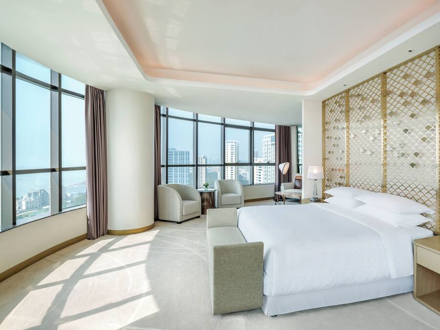 A hotel room with a large bed, a seating area, a desk, and a view of the cityscape through large windows.