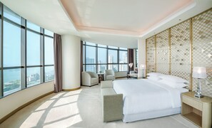 A hotel room with a large bed, a seating area, a desk, and a view of the cityscape through large windows.