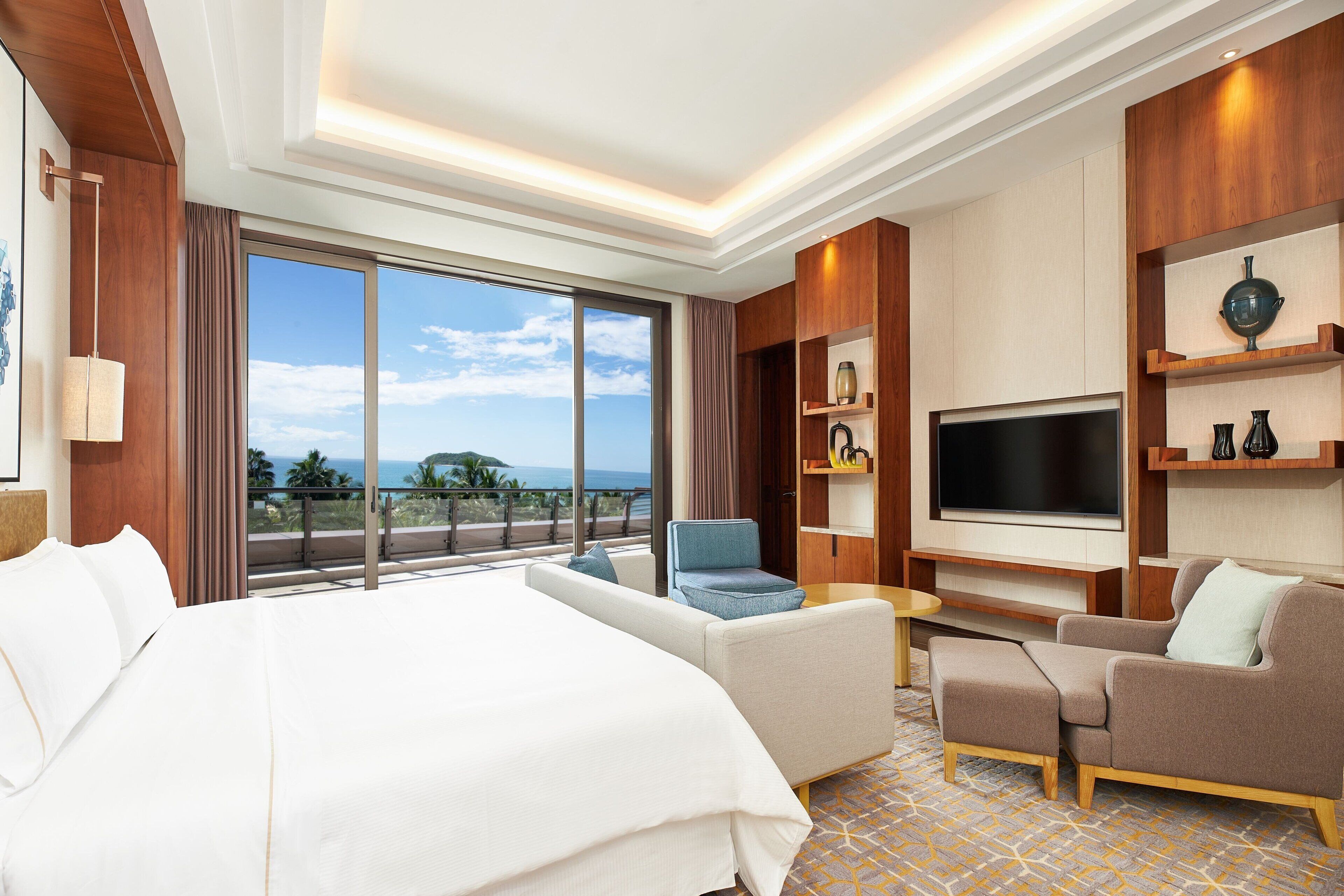A hotel room with a large bed, a sofa, a TV, and a balcony with a view of the ocean.