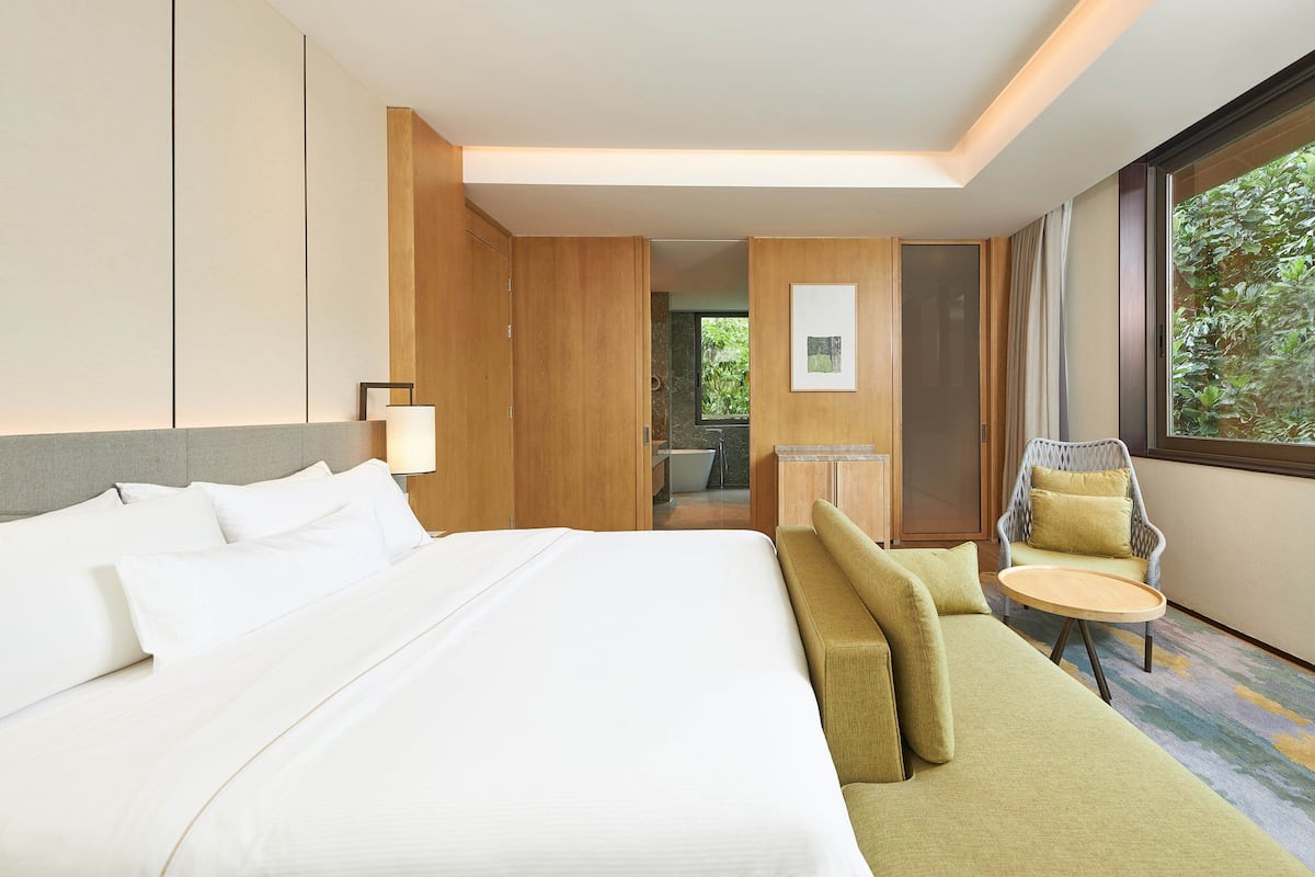 A hotel room with a large bed, a sofa, a small table, and a chair. There is a window with a view of greenery outside.