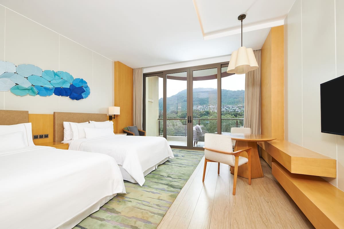 A hotel room with two beds, a desk, a chair, a TV, and a large window with a view of a city and mountains.