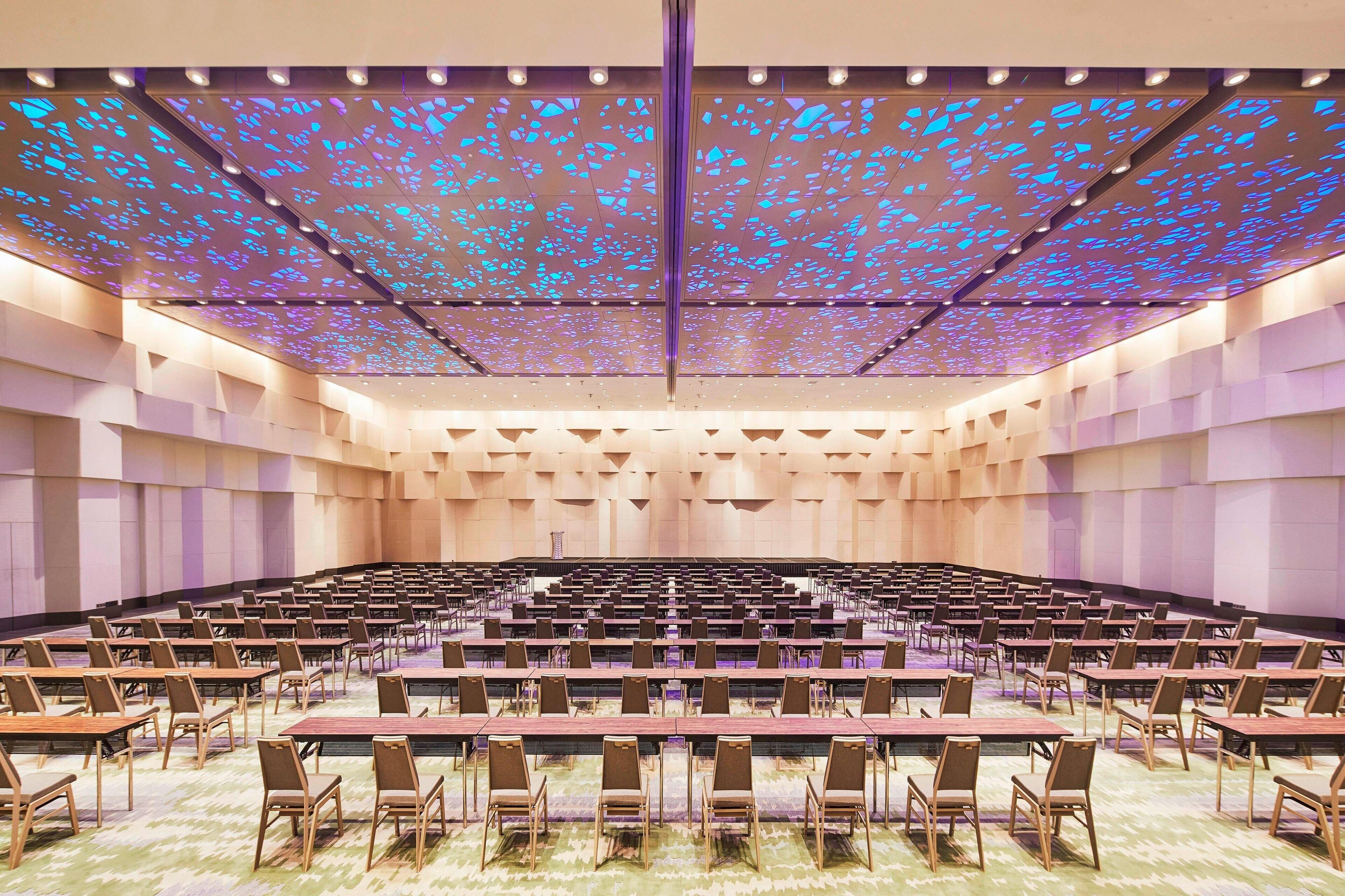 Un salón de conferencias con filas de mesas y sillas, un techo alto con un diseño de luces decorativo y un escenario central.