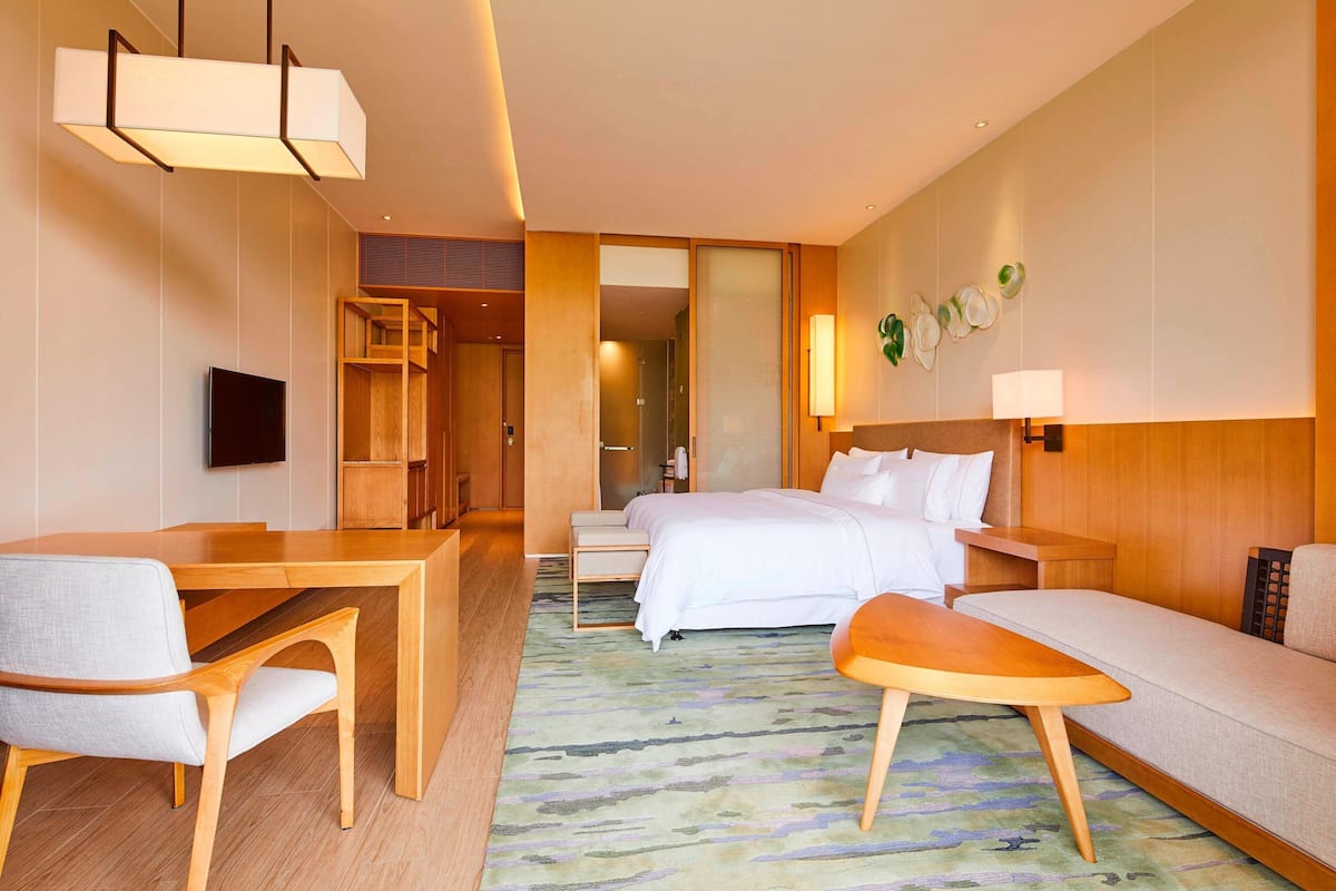 A hotel room with a bed, a desk, a chair, a sofa, a TV, and a wall-mounted lamp.