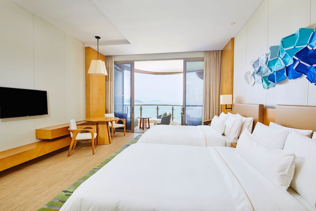 A hotel room with two beds, a desk, a chair, a TV, and a large window with a view.