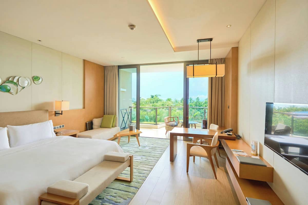 A modern hotel room with a large bed, a desk, a chair, a sofa, and a balcony with a view.