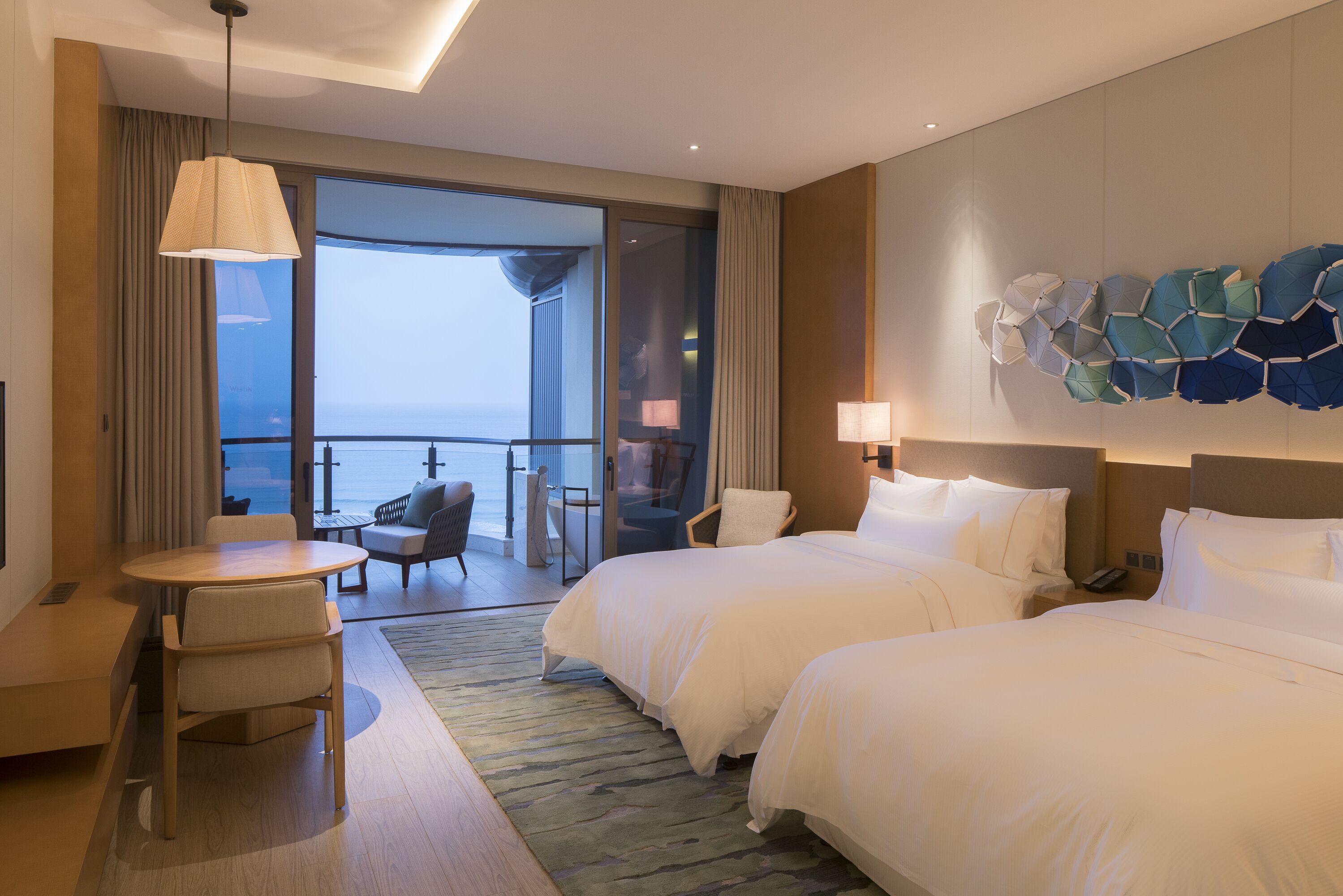 A hotel room with two beds, a seating area with a table and chairs, a large window with a view of the sea, and a decorative wall piece.