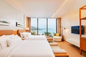 Two large beds with white linens, a flat-screen TV, and a sofa in a room with a sea view.