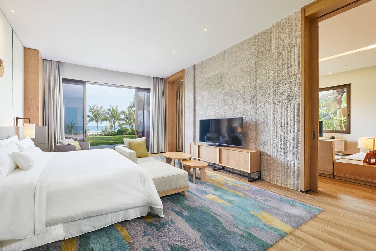 A modern hotel room with a large bed, a flat-screen TV, a small seating area, and a view of palm trees through large windows.