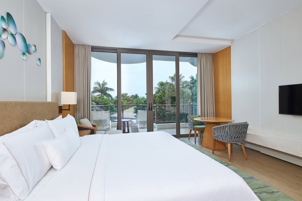A hotel room with a large bed, a desk with a chair, a TV, and a balcony with a view of palm trees.