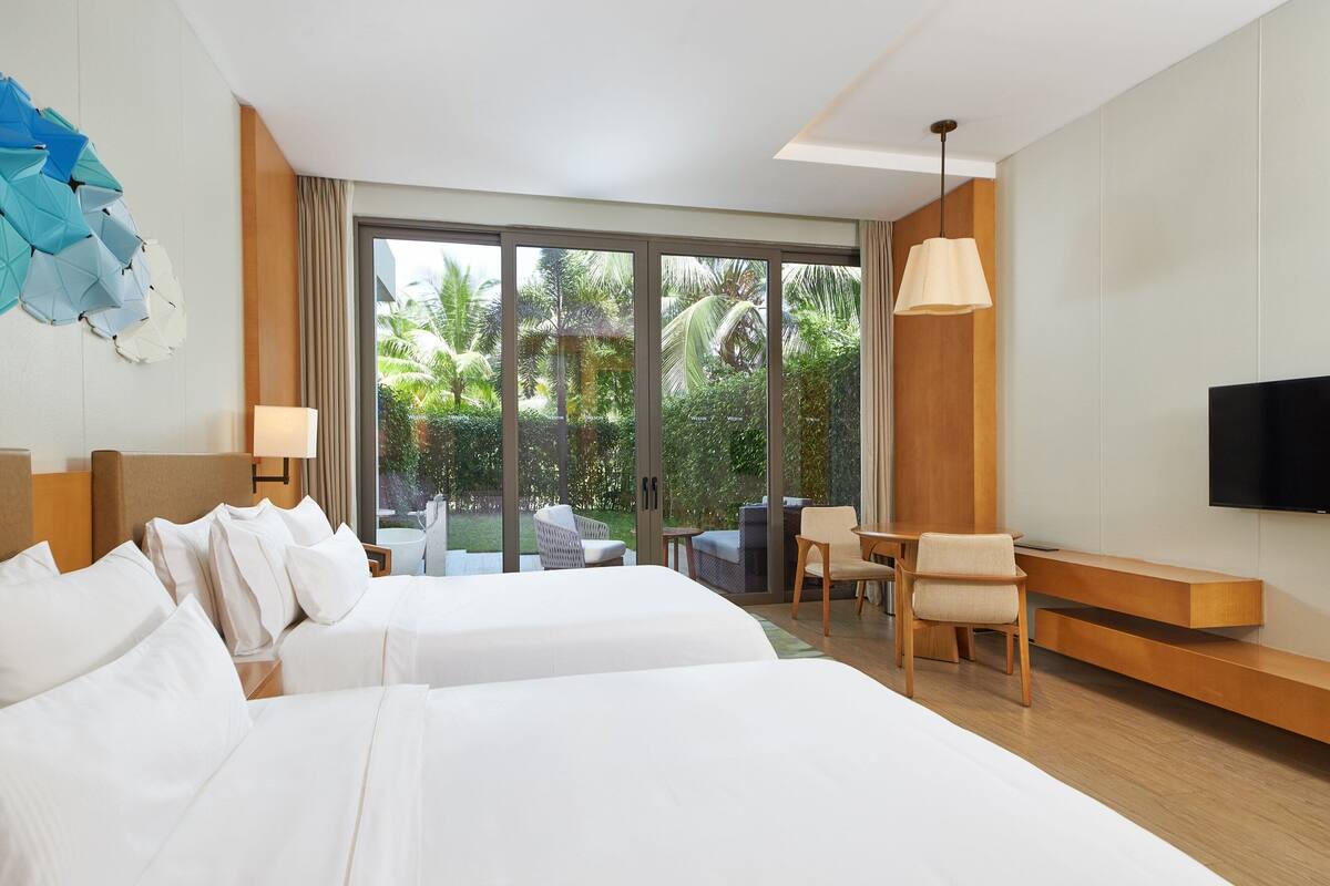 A hotel room with two beds, a TV, a seating area with a table and chairs, and a large window offering a view of greenery.