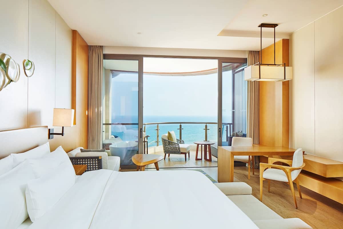 A modern hotel room with a large bed, a desk, and a balcony with a view of the sea.