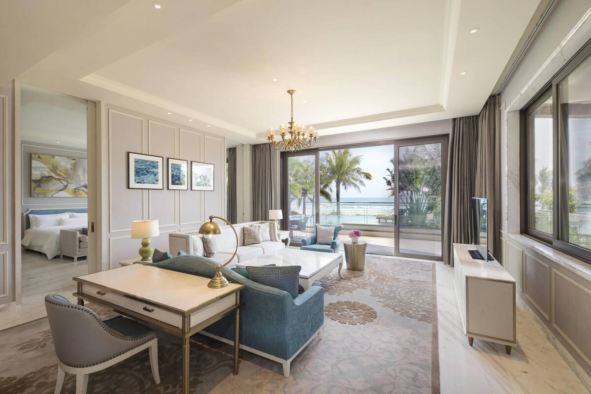 A hotel room with a large window, a dining area, a sitting area with a sofa and armchairs, and a desk with a lamp.
