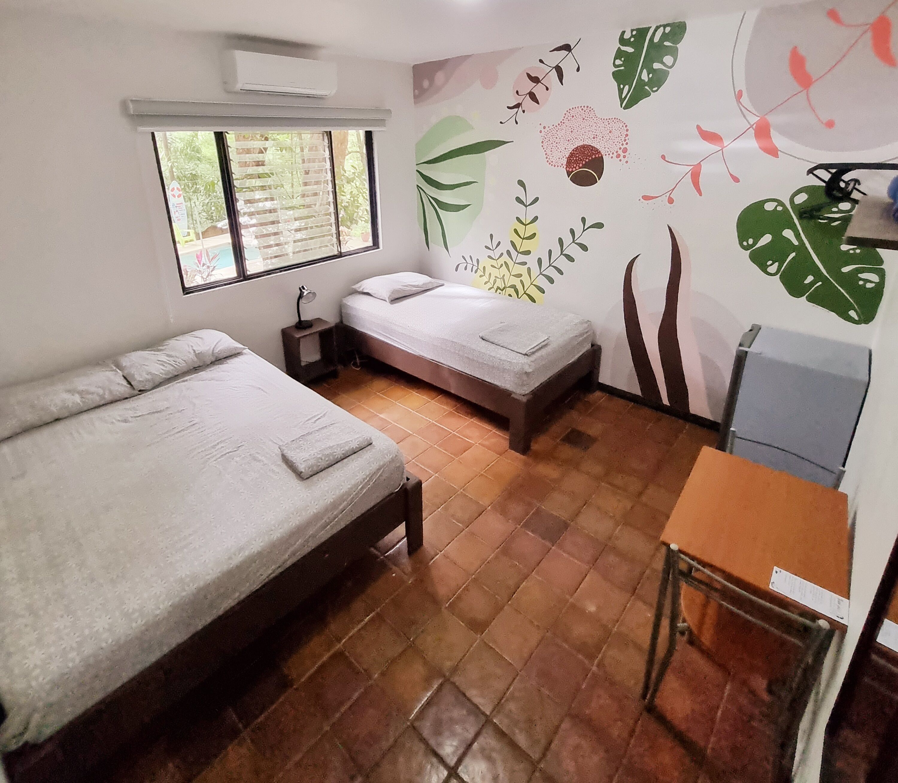 A small, clean room with two single beds, a desk, and a large window.