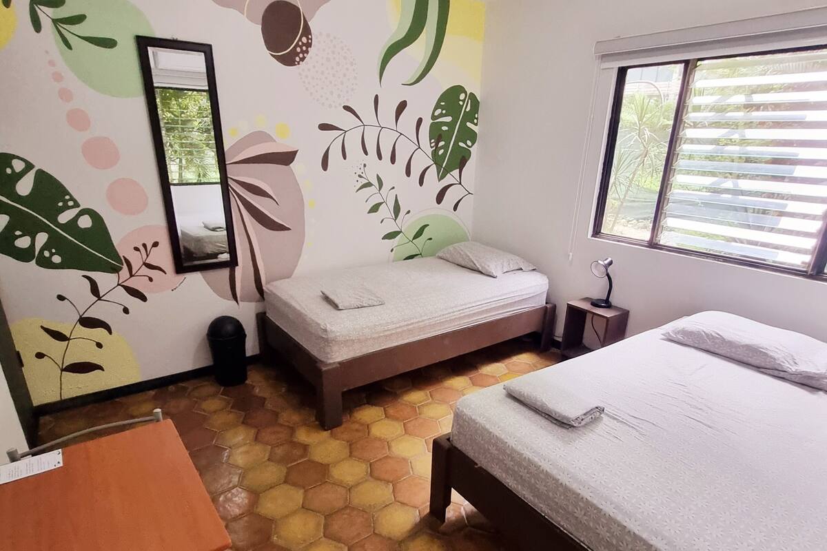 Une chambre avec deux lits, un bureau et une chaise. Les murs sont décorés d’un motif de feuilles.