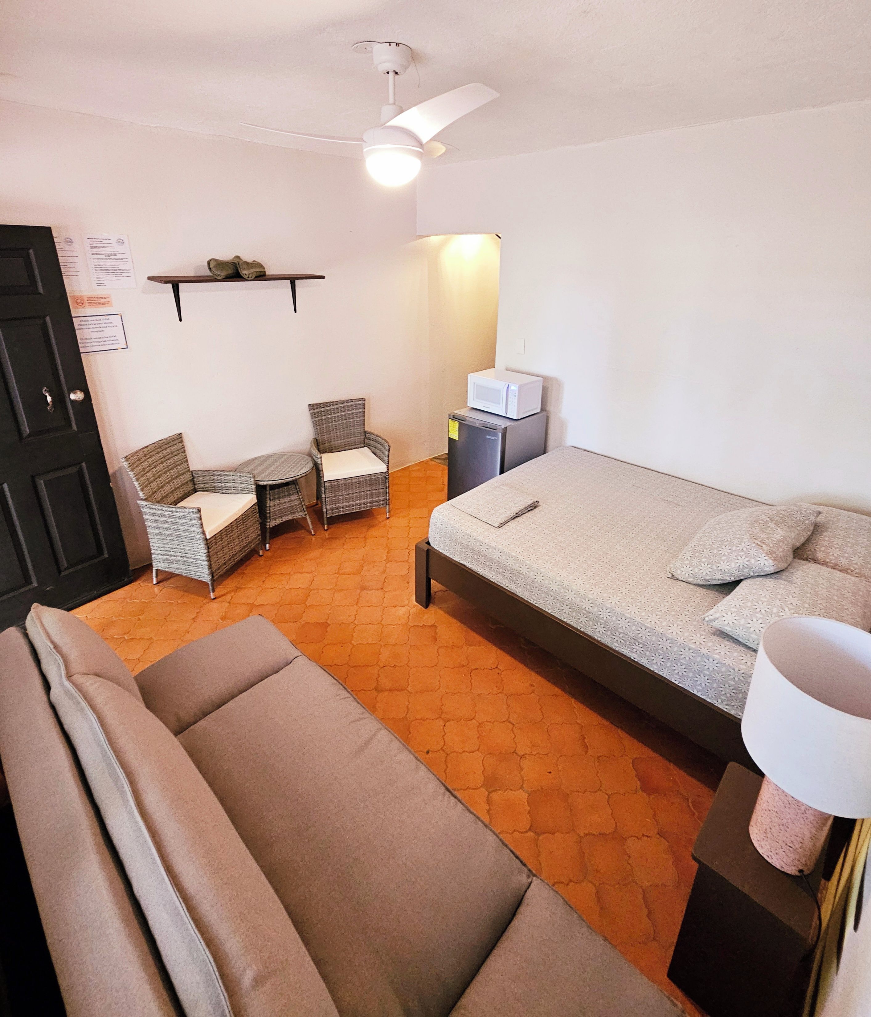 Pequeña habitación amoblada con cama, sofá, dos sillas, microondas y un basurero.