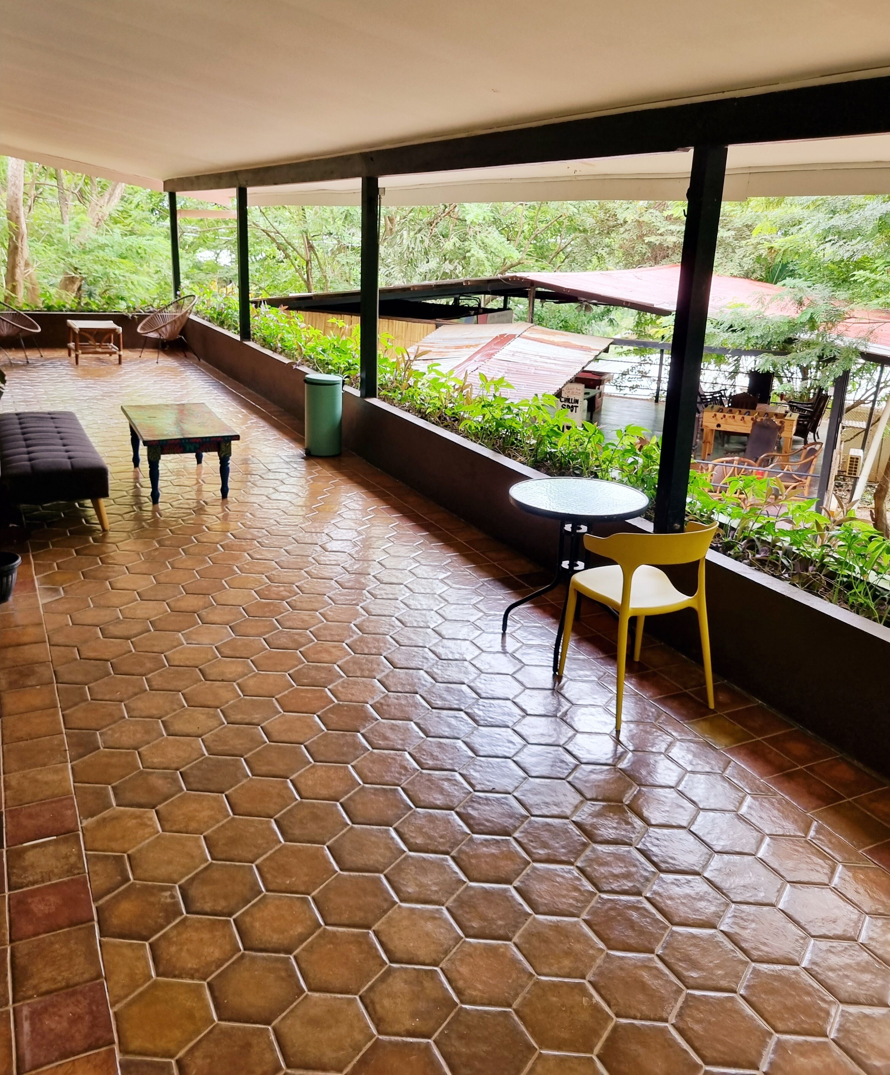 Een overdekt terras met hexagonale tegels, buitenmeubels waaronder een bank en tafels, en een uitzicht op groen en een gebouw op de achtergrond.