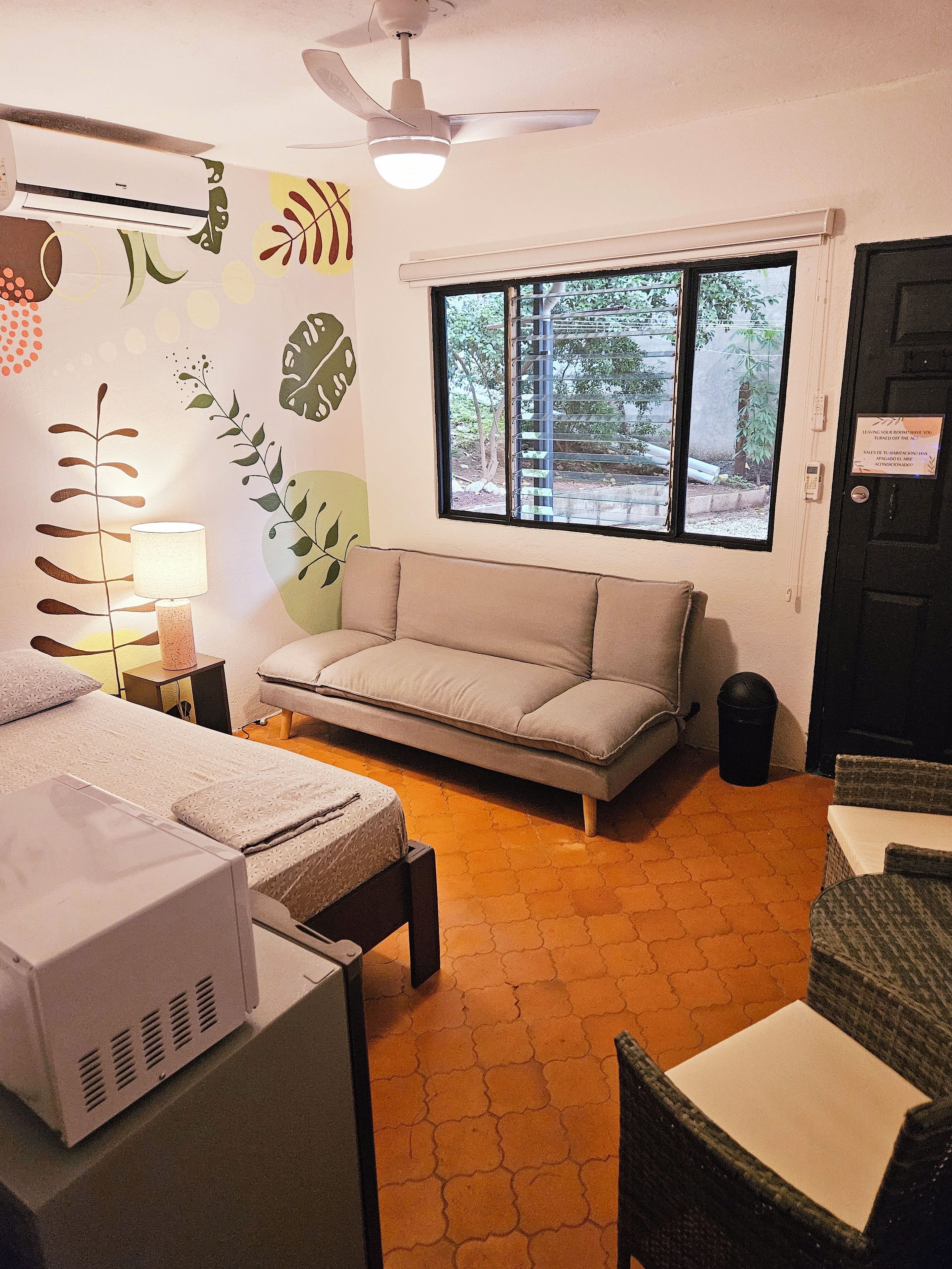 A compact room with a bed, a sofa, a table, a lamp, a ceiling fan, and a window with a view of greenery.