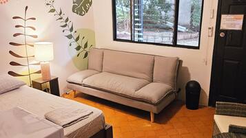 Een kleine kamer met een bed, een bank, een tafel, een lamp, een plafondventilator en een raam met uitzicht op groen.