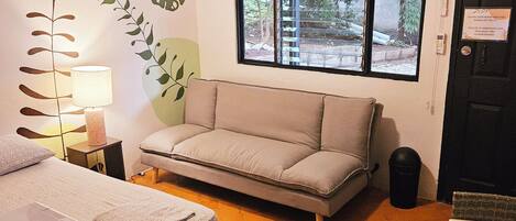 A compact room with a bed, a sofa, a table, a lamp, a ceiling fan, and a window with a view of greenery.