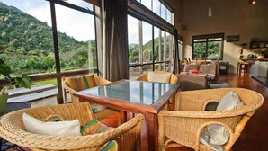 A dining area with wicker furniture, large windows, and a view of a mountainous landscape. - Piha Beachstay - Hostel (Piha)