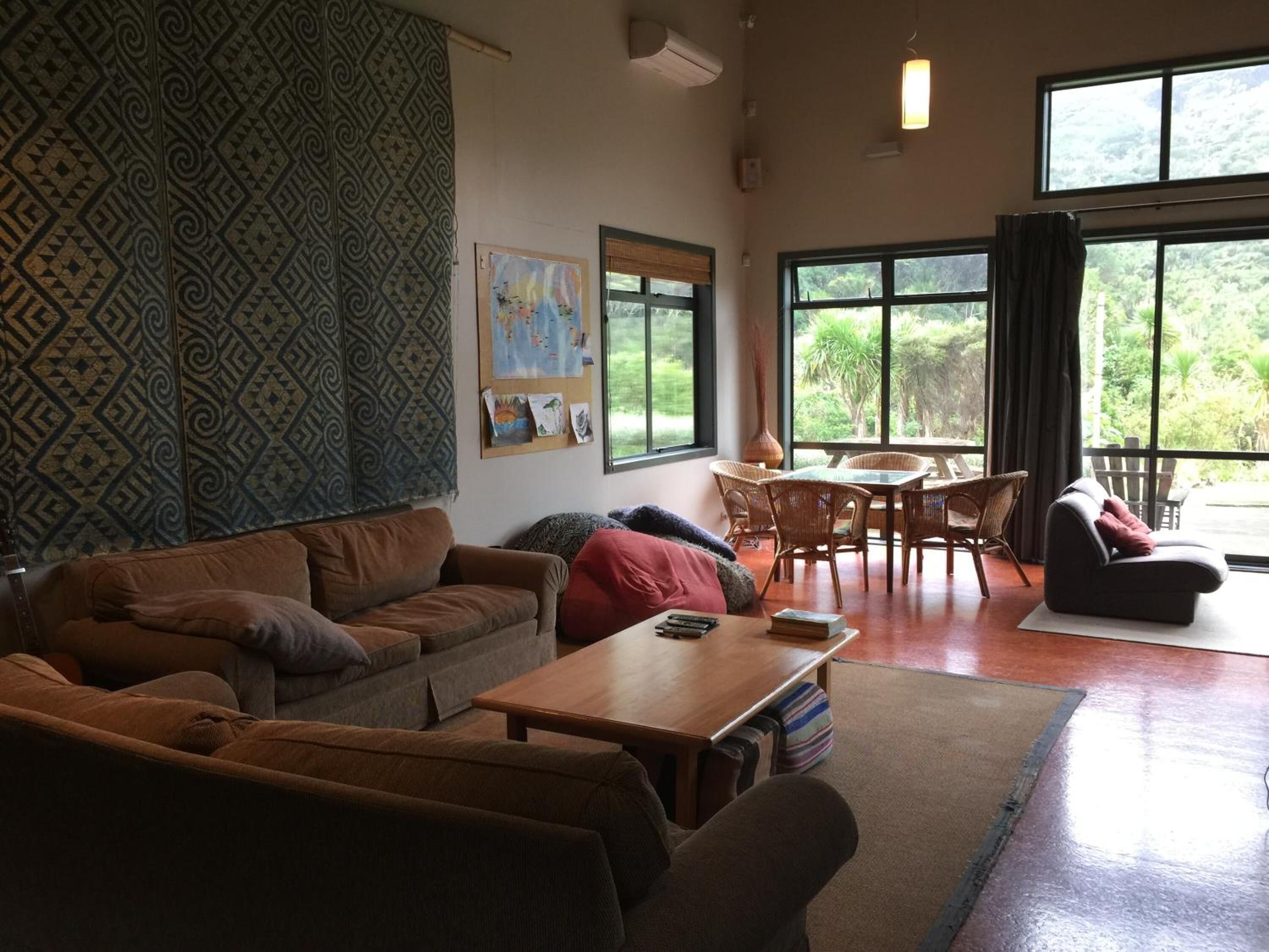 Een ruime woonruimte met een behang, een houten bijzettafeltje, een bank, een eettafel met stoelen, en grote ramen met uitzicht op groen.