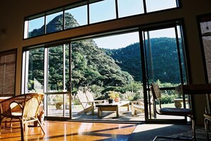 A spacious room with large windows offering a view of a lush forested mountain. - Piha Beachstay - Hostel (Piha)