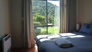 A bedroom with a large window, a bed with a white towel, and a view of a green landscape. - Piha Beachstay - Hostel (Piha)