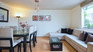 A modern living room with a glass dining table, a sofa, and a coffee table. There are three framed artworks on the wall and a vase with flowers on the table. - Duplex Apartment in Miraflores (Lima)