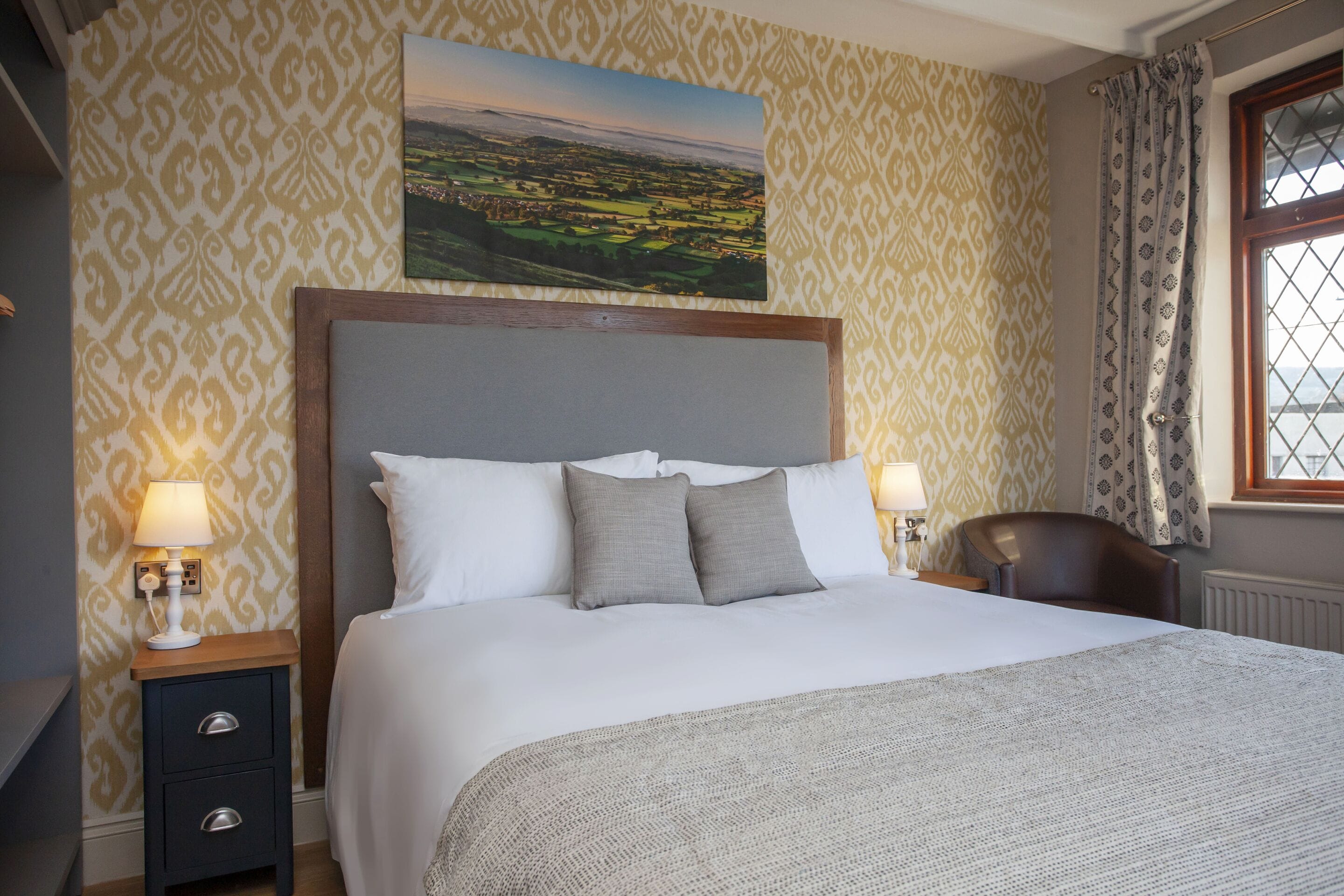 A neatly made bed with a headboard, two bedside tables with lamps, a chair, and a window with curtains.
