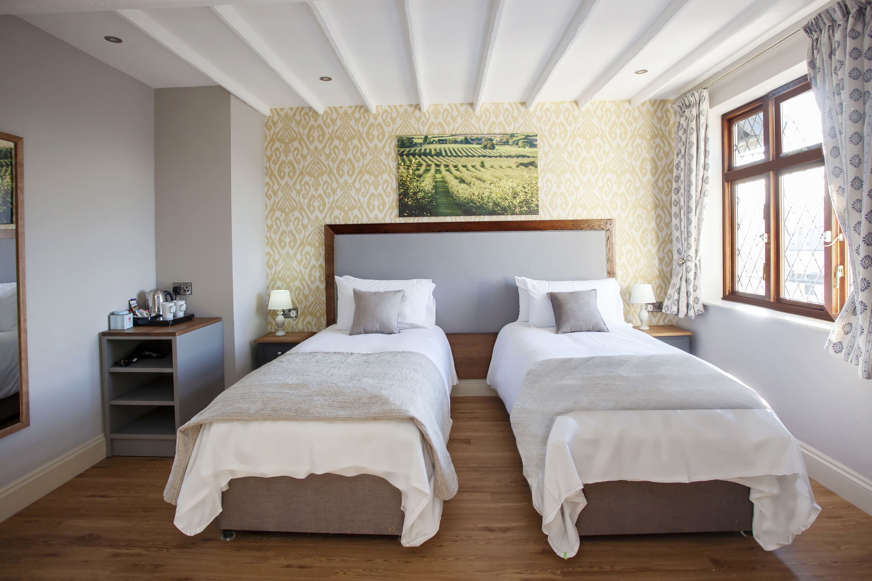 A double room with two beds, a wooden floor, a patterned wall, and a window with curtains.