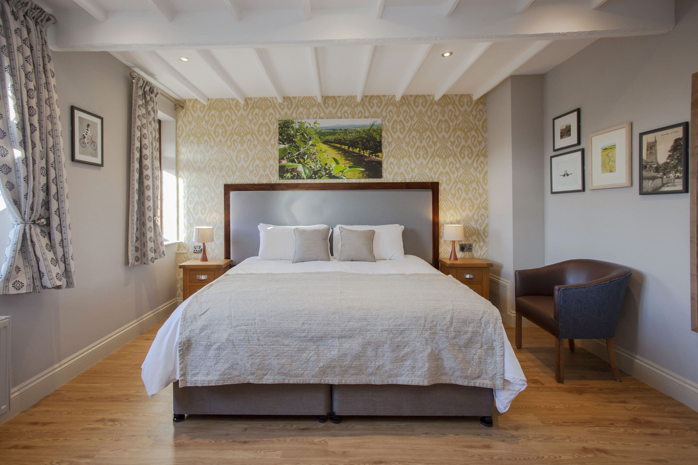 Ein geräumiges Schlafzimmer mit einem großen Bett, Holzboden, einer Kopfteil mit Landschaftsbild und einem Sessel. An der Wand befinden sich gerahmte Bilder.