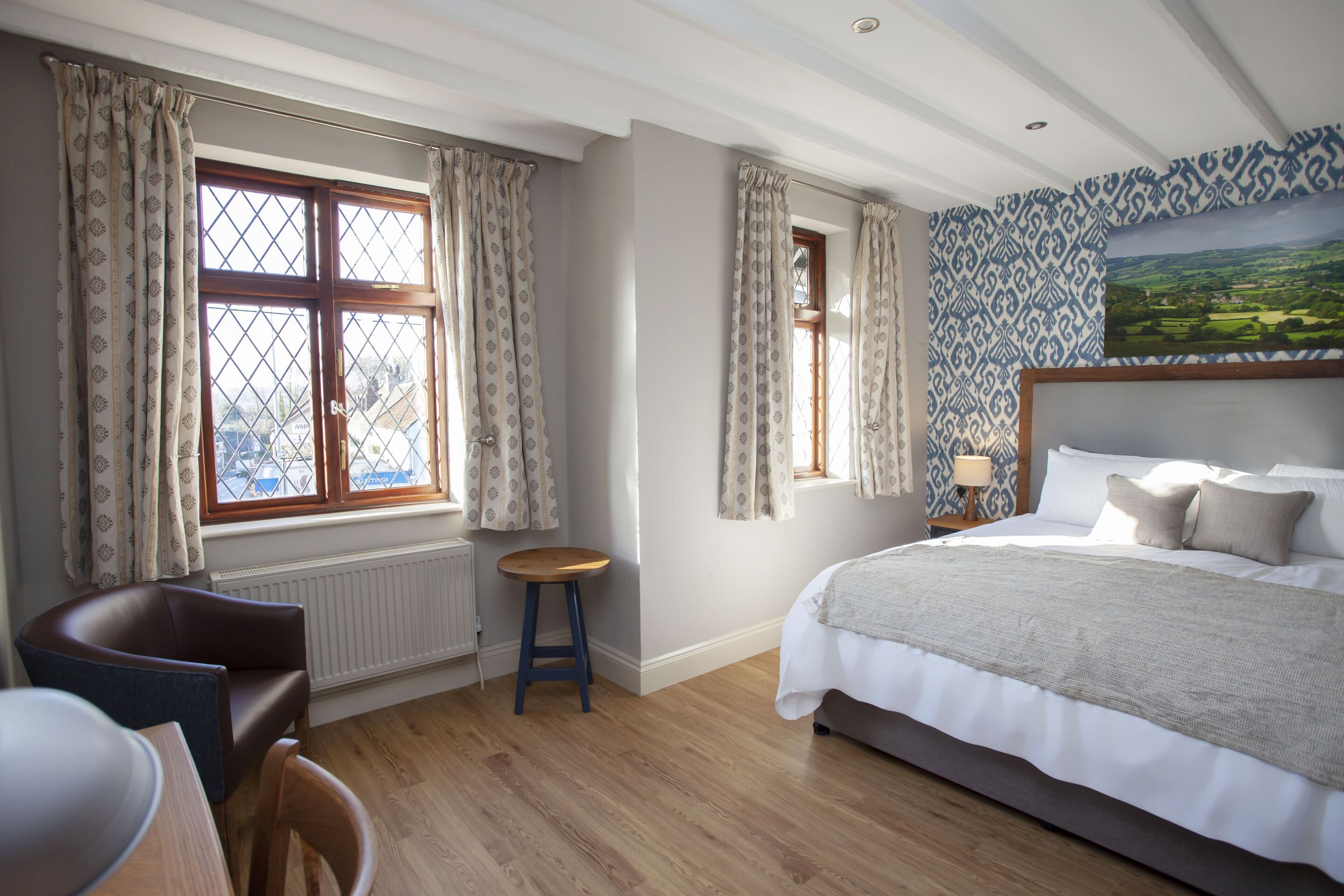 A bedroom with a large bed, a chair, a small stool, and two windows with curtains.