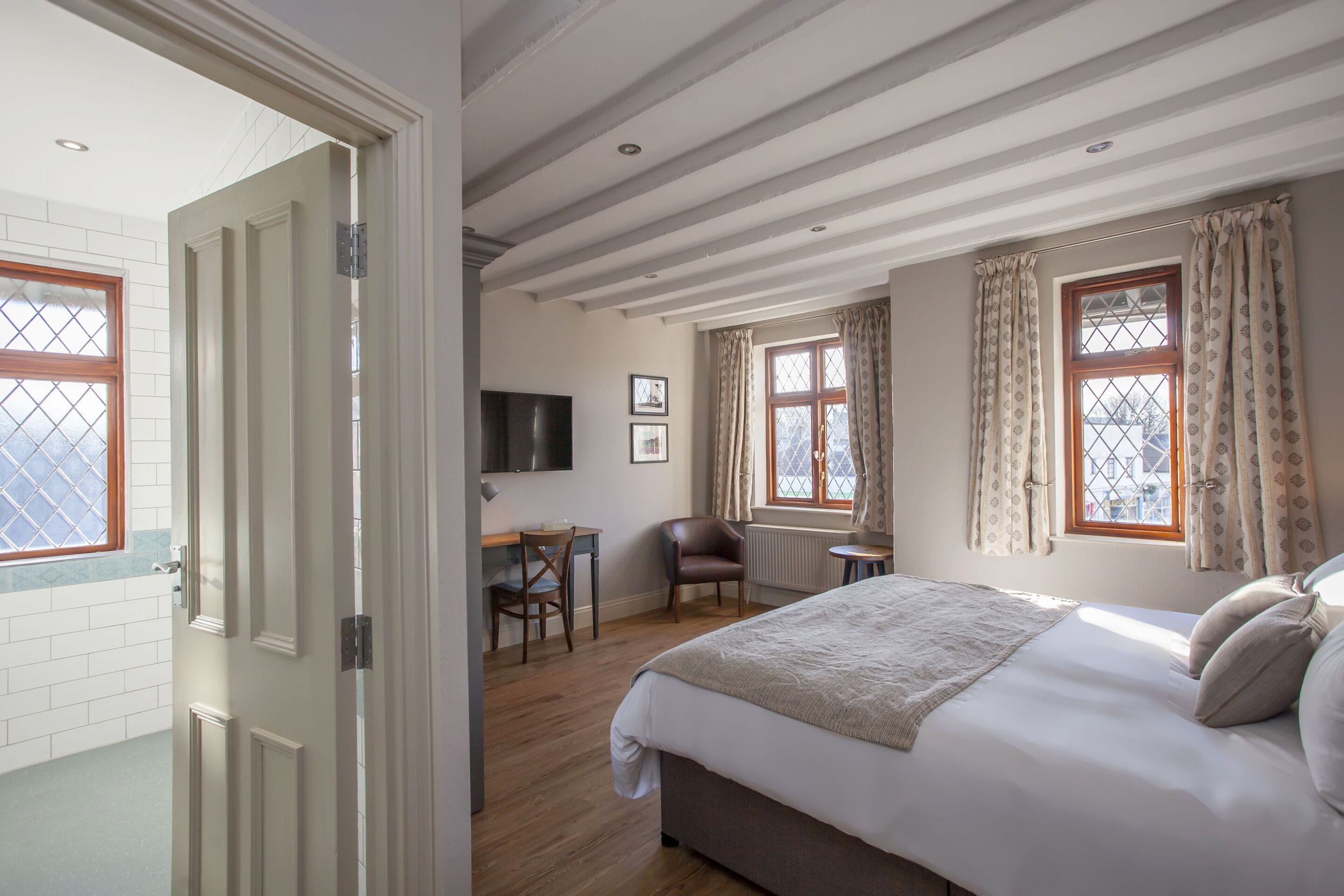 A hotel room with a large bed, a desk, a chair, and two windows with curtains.