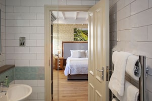 A hotel room with a wooden floor, a bed with white bedding, a nightstand, a wall-mounted towel rack, and a bathroom with a sink and mirror.