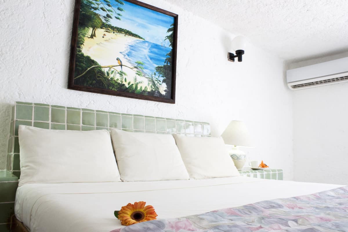A hotel room with a bed, a painting of a beach scene, a nightlight, and an air conditioning unit.