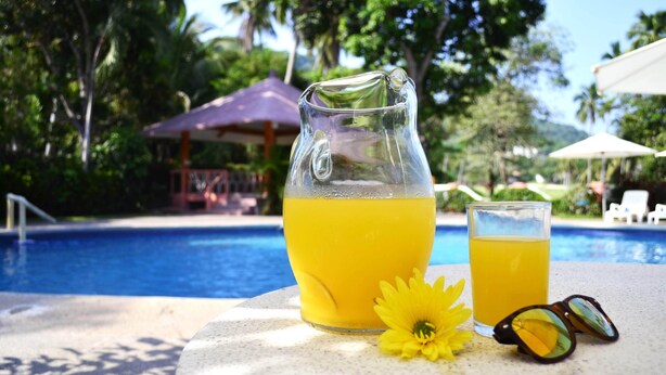 A glass of orange juice and a pitcher on a table by a pool with umbrellas and palm trees in the background. - Coral Ixtapa (Ixtapa)