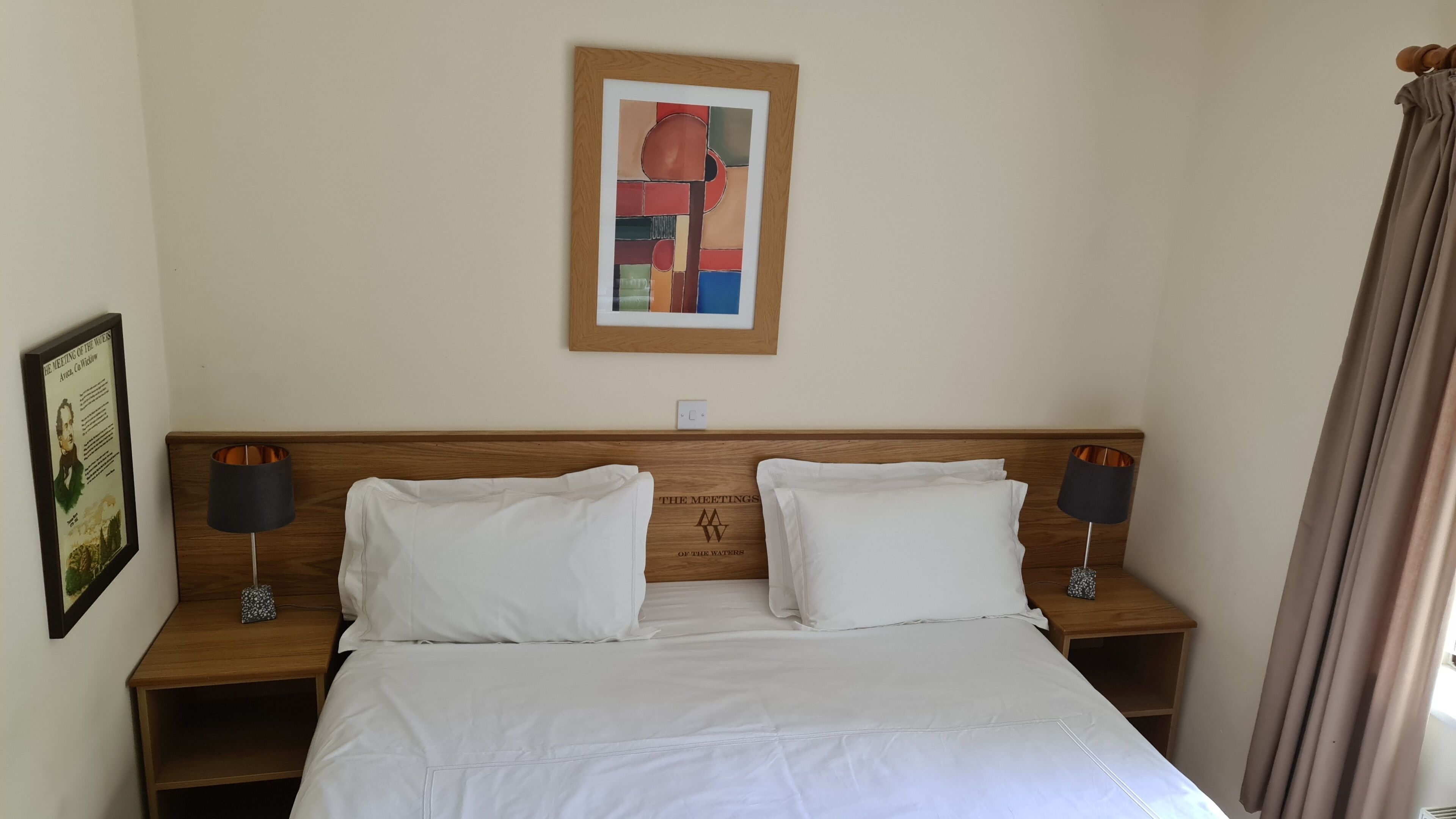 A neatly made bed with white linens, two bedside tables with lamps, a framed abstract painting above the bed, and a framed informational poster on the wall.