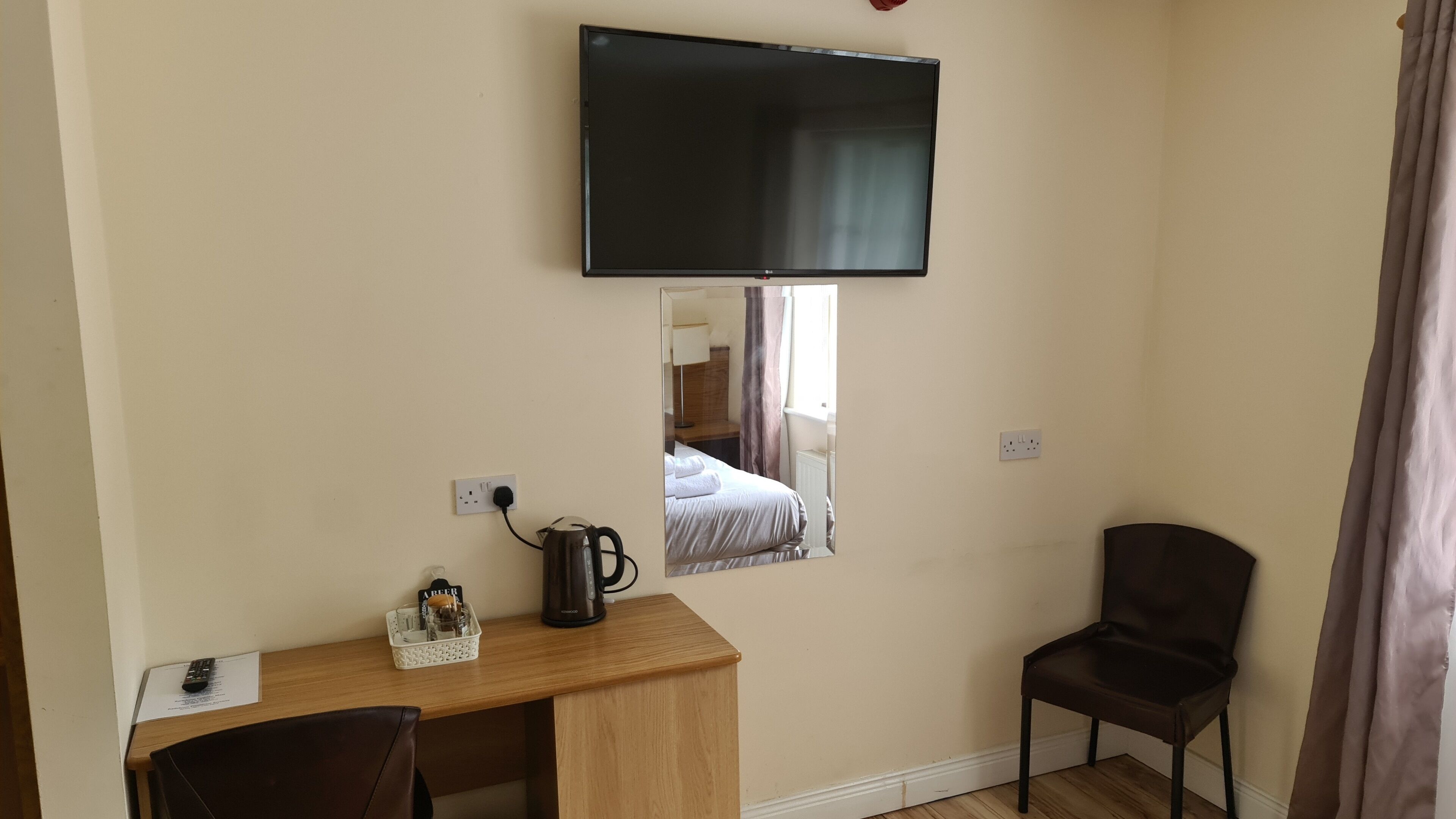 A hotel room with a wooden desk, a chair, a flat-screen TV mounted on the wall, and a bed with a mirror reflecting the room.