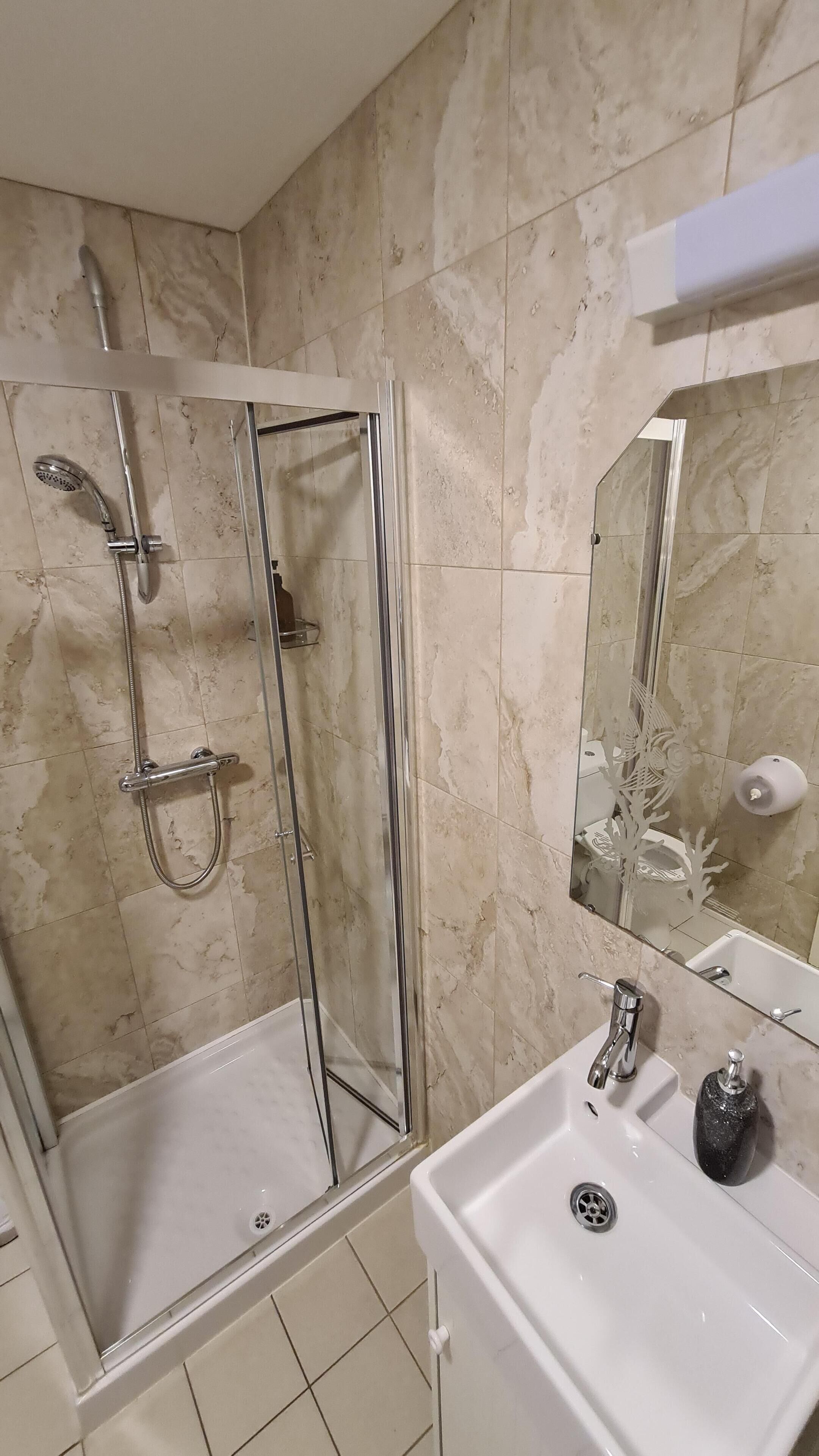 Un baño con ducha de vidrio, un lavamanos blanco con dispensador de jabón negro y un espejo sobre el lavamanos.